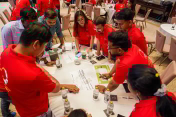 Team members in red shirts participating in a hands-on team-building activity around a table, collaborating on a group problem-solving exercise.